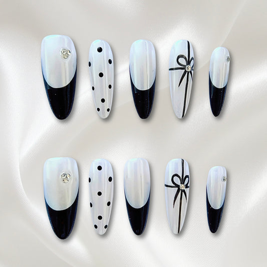 A set of press-on nails showcasing various designs including black French tips, rhinestones, polka dots, and bow charms on a pearl-white base.